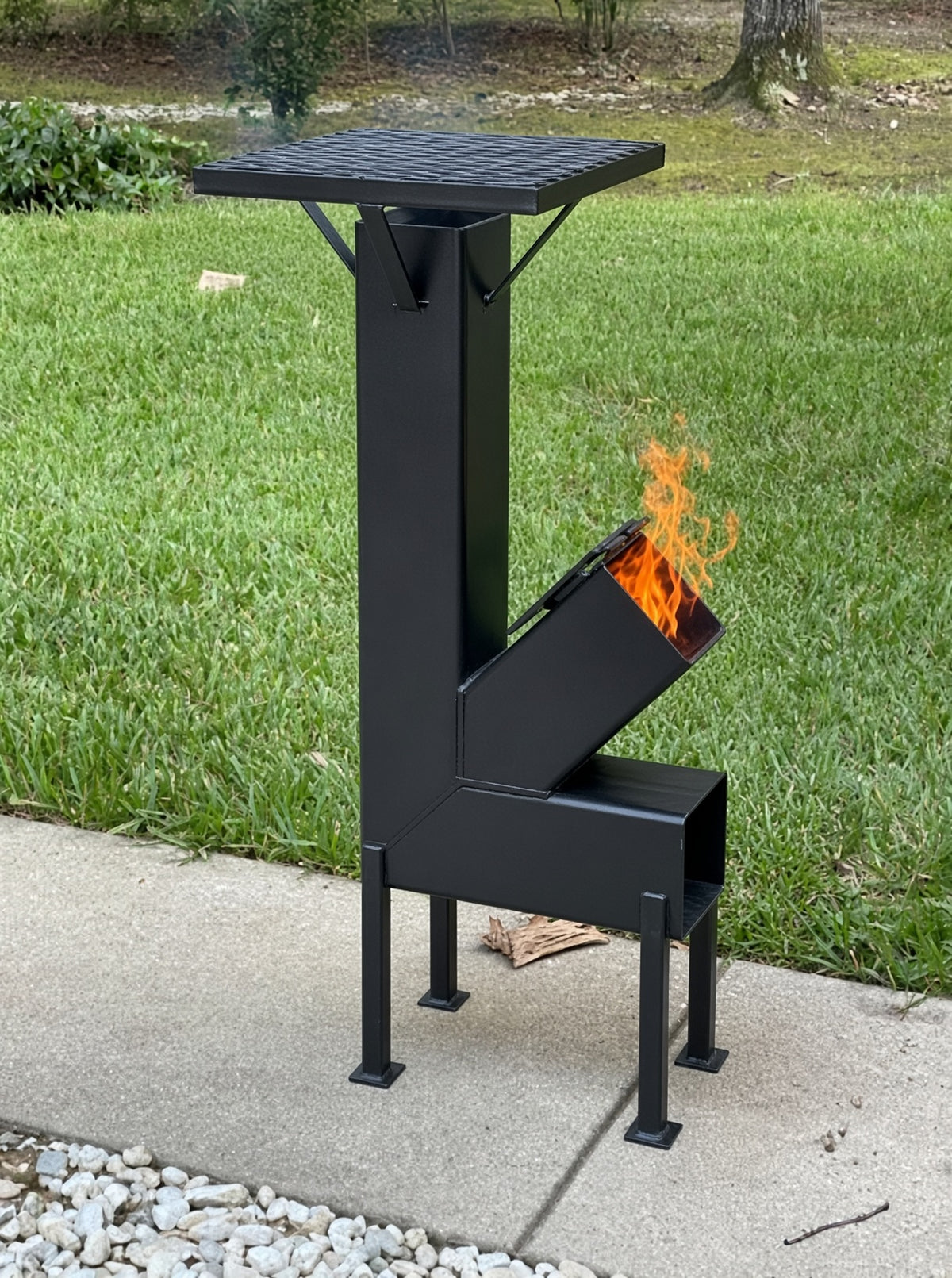Steel rocket stove burning kindling outdoors on a backyard patio, ready for off-grid cooking